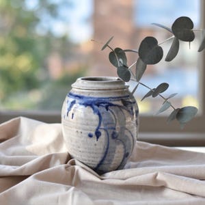 May include: A handmade ceramic vase with a lid, showcasing a white base and blue decorative patterns. The vase is placed on a beige cloth, with a sprig of eucalyptus. The vase is approximately 15 cm tall.