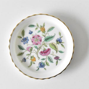Könnte beinhalten: Kleiner, weißer Keramikteller mit goldenem Rand und floralem Design. Das Design zeigt rosa, blaue und grüne Blumen und Blätter. Der Teller hat einen gewellten Rand.