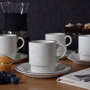 Op de afbeelding: Een set van gespikkelde witte keramische mokken en schotels met een donkere rand. De mokken zijn op de schotels gestapeld. Een glazen koffiekan, muffins, blauwe bessen en een servet met bestek zijn ook zichtbaar.