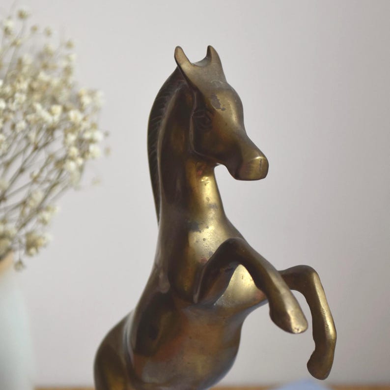 May include: A brass horse figurine depicted in a rearing pose. The sculpture features a smooth, metallic surface with a warm, golden-brown patina, showing the horse with its front legs lifted and head turned slightly to the right side.