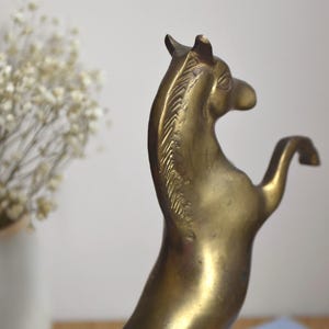 May include: A brass horse figurine in a rearing position, showing a smooth metallic surface with a golden-brown tone. The sculpture features detailed etched lines along the mane and a curved neck, capturing the animal in a dynamic pose.