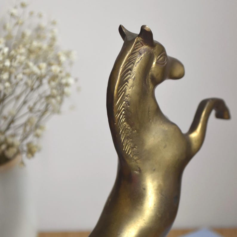 May include: A brass horse figurine in a rearing position, showing a smooth metallic surface with a golden-brown tone. The sculpture features detailed etched lines along the mane and a curved neck, capturing the animal in a dynamic pose.