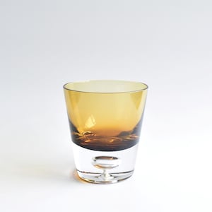 Op de afbeelding: Een helder glas met een uniek ontwerp. Het bovenste gedeelte is amberkleurig, overgaand in een donkerder bruin aan de basis. Het onderste gedeelte is helder, met een kleine, afgeronde basis. Geschikt voor dranken.