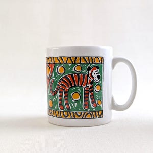 Op de afbeelding: Witte keramische mok met een tijgerillustratie. De tijger heeft oranje en zwarte strepen op een groene achtergrond met gele cirkels. De mok heeft een gebogen handvat.