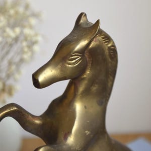 May include: A brass horse figurine in a rearing pose, featuring a polished metallic gold finish with etched details on the eyes and mane. The sculpture captures the animal with its front legs lifted, showcasing a smooth, reflective surface.