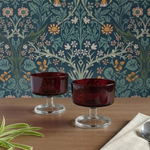Op de afbeelding: Twee robijnrode glazen dessertkommen met heldere stelen en bodems. De kommen staan op een houten oppervlak met een bloemenbehang achtergrond. Een lepel en een servet bevinden zich aan de rechterkant van de afbeelding.