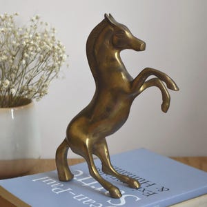 May include: A brass horse figurine depicted in a rearing pose with its front legs lifted. The sculpture features a smooth, metallic gold-toned surface with subtle variations in color, showcasing a stylized mane and defined facial features.