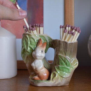May include: A ceramic match holder with a bunny figurine and a tree stump design. The holder is filled with matches. The bunny has orange and white fur, and the holder has green leaves and a brown tree trunk.