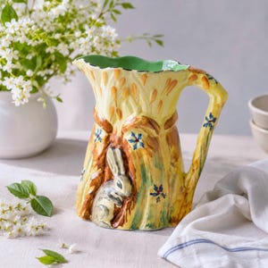 Op de afbeelding: Handbeschilderde keramische kan met een grillig ontwerp. De kan heeft een geel en bruin boomstammotief met een konijn erin genesteld. Blauwe bloemaccenten en een groene rand maken het ontwerp compleet. Een decoratief stuk voor in huis.