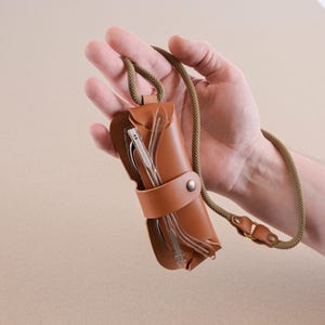 May include: A brown leather glasses case with a strap. The case is closed and has a pair of glasses inside. The strap is made of a brown cord.