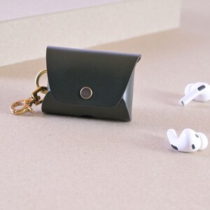 Könnte beinhalten: Dunkelgrüne Leder-AirPod-Hülle mit goldfarbenem Verschluss und Knopfverschluss. Zwei weiße kabellose Ohrhörer liegen neben der Hülle auf einer neutralen Oberfläche. Die Hülle dient zum Schutz und Transport kleiner elektronischer Geräte.