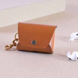 Könnte beinhalten: Eine braune Leder-AirPod-Hülle mit einem goldfarbenen Verschluss und Knopfverschluss. Die Hülle ist rechteckig mit abgerundeten Kanten. Zwei weiße AirPod-Ohrhörer sind im Hintergrund zu sehen.