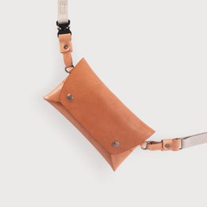 May include: A tan leather fanny pack with a white and black adjustable strap. The bag has a flap closure with a snap button.