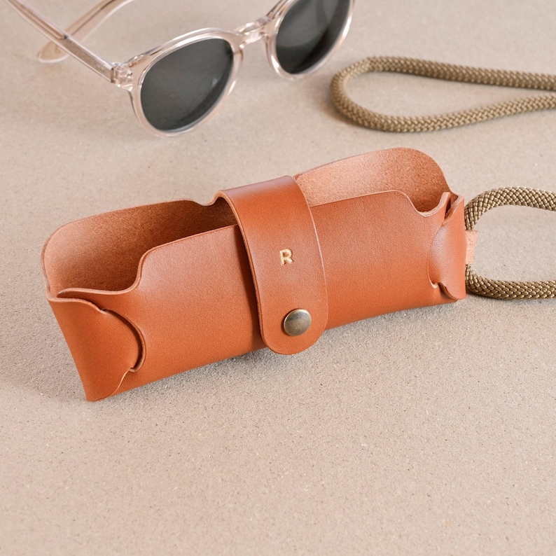 May include: A brown leather glasses case with a strap and a metal button. The case has the letter "R" embossed on the front.