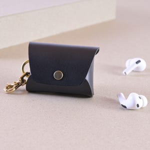 May include: A black leather earbud case with a gold-coloured clasp and button closure. Two white wireless earbuds are next to the case. The case is designed to hold and protect small electronic devices.