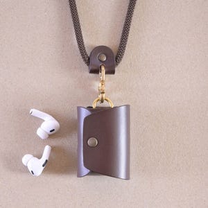 Könnte beinhalten: Eine braune Leder-AirPod-Hülle mit Druckknopfverschluss, befestigt an einer dunkelgrauen Kordelkette. Die Hülle hat einen goldfarbenen Verschluss. Zwei weiße kabellose Ohrhörer liegen neben der Hülle.