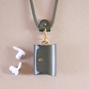 Könnte beinhalten: Olivgrünes Leder-AirPod-Etui mit goldfarbenem Verschluss und passendem grünem Kordel. Zwei weiße kabellose Ohrhörer liegen neben dem Etui. Das Etui hat einen Druckknopfverschluss und eine Schlaufe für die Kordel.