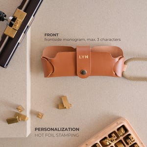 May include: A brown leather glasses case with a gold clasp and the name "LYN" embossed on the front. The case is shown on a light brown surface with gold metal letters scattered around it. The text "FRONT" and "frontside monogram, max. 3 characters" is above the case. The text "PERSONALIZATION HOT FOIL STAMPING" is below the case.