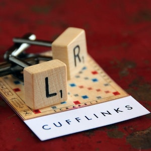 May include: A pair of cufflinks made from wooden Scrabble tiles, one with the letter "L" and the other with the letter "R". The cufflinks are displayed on a white card with the word "CUFFLINKS" printed on it.