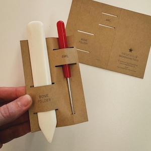 Può includere: Un kit di strumenti in cartone marrone con un piegatore per ossa, un punteruolo e un'etichetta che recita "Shelbyville Bookbinding Tools".