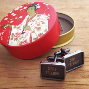 May include: A pair of silver cufflinks with black accents. The cufflinks have the words "Shift Freedom" engraved on them. The cufflinks are sitting on a wooden surface next to a red and gold round box with a floral design.