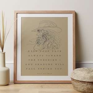 Puede incluir: Una impresión enmarcada con un fondo beige que presenta un dibujo lineal de un hombre con barba y sombrero. La cita "Keep your face always toward the sunshine - and shadows will fall behind you. Walt Whitman" está impresa debajo del dibujo.