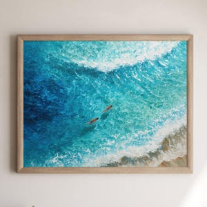 May include: An aerial view painting of two people swimming in turquoise ocean water. The artwork is framed in a light brown wooden frame. The ocean waves are white and the sand is light brown.