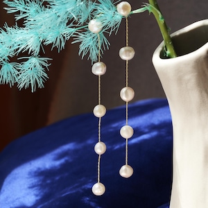 Könnte beinhalten: Ein Paar lange, zarte goldene Kettenohrringe mit weißen Perlenakzenten. Die Ohrringe hängen an einer Vase mit blauem Hintergrund.
