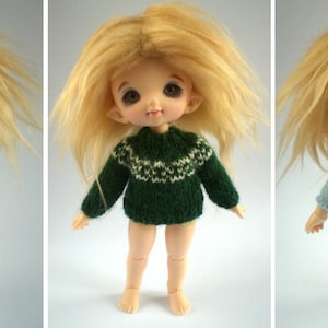May include: Three dolls with blonde hair and large eyes are displayed. Each doll wears a different knitted outfit: a green cardigan with cream pants, a green sweater with a white pattern, and a blue wrap sweater with cream pants. The dolls are on a white background.