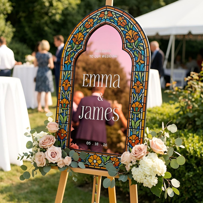May include: An arched wedding welcome sign with a stained glass border featuring orange and blue floral patterns. The centre is a reflective surface with the white text: WELCOME TO OUR WEDDING, EMMA & JAMES, 05.18.26. It is decorated with pink roses and eucalyptus.