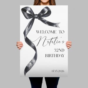 May include: A rectangular white sign featuring a large, hand-drawn style black ribbon bow graphic on the left side. The text reads: WELCOME TO Natalie's 32ND BIRTHDAY 07.15.2026. The design uses a mix of bold sans-serif and elegant script typography.