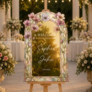 May include: An arched wedding welcome sign featuring a stained glass border with pink floral motifs and vines. The centre displays the text: WELCOME TO THE WEDDING OF Sophie AND John 07.14.2026. The sign is mounted on a wooden easel stand.