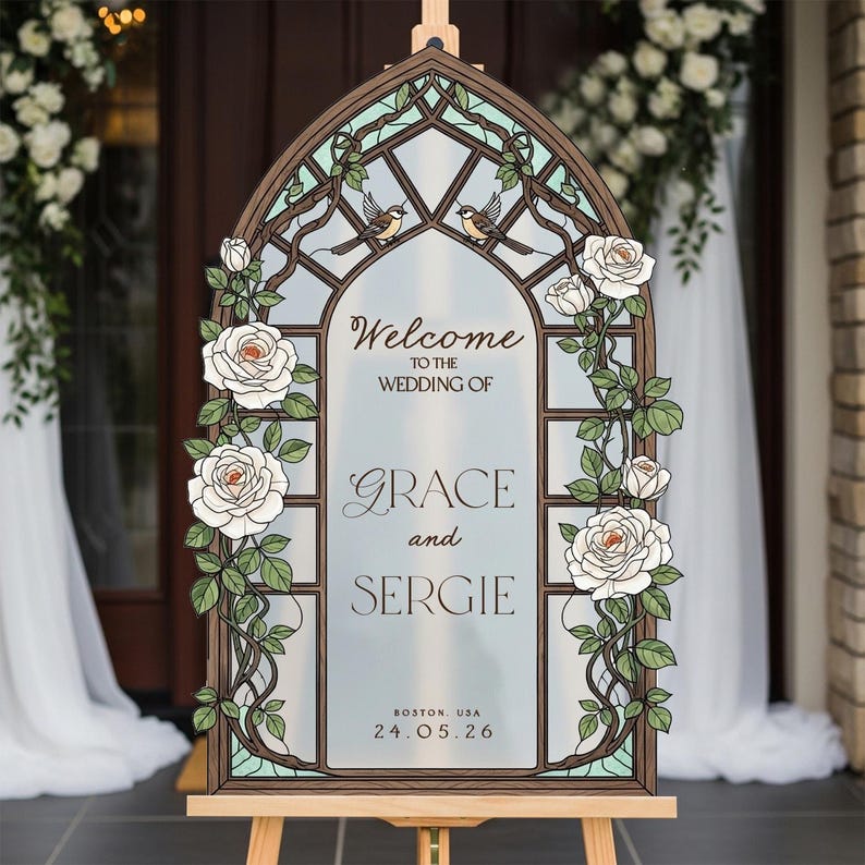 May include: An arched wedding welcome sign featuring a stained glass window design with brown frames, white roses, green leaves, and two small birds. The text reads: Welcome to the wedding of Grace and Sergie, Boston, USA, 24.05.26, printed on a frosted surface.