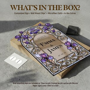 May include: A rectangular wedding sign featuring a stained glass-style border with purple iris flowers and gold scrollwork. The center text reads "WELCOME TO OUR WEDDING EMMA & JAMES 07.08.26". The sign rests on a cardboard box with a small white card and a black pouch.