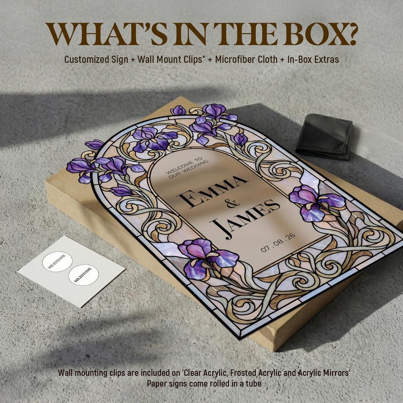 May include: A rectangular wedding sign featuring a stained glass-style border with purple iris flowers and gold scrollwork. The center text reads "WELCOME TO OUR WEDDING EMMA & JAMES 07.08.26". The sign rests on a cardboard box with a small white card and a black pouch.
