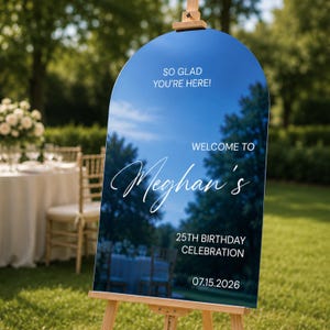 May include: An arched, reflective welcome sign on a wooden easel. The sign features white text that reads: SO GLAD YOU'RE HERE! WELCOME TO Meghan's 25TH BIRTHDAY CELEBRATION 07.15.2026. The surface reflects the surrounding outdoor environment.