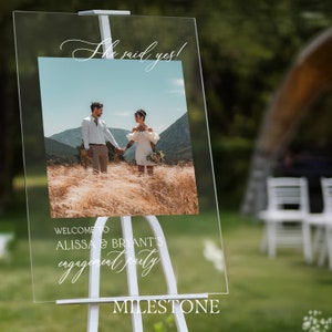 May include: A clear acrylic rectangular sign mounted on a white easel. The sign features the text "She said yes!" at the top, a central photograph of a couple in a field, and the text "WELCOME TO ALISSA & BRYANT'S engagement party" below.