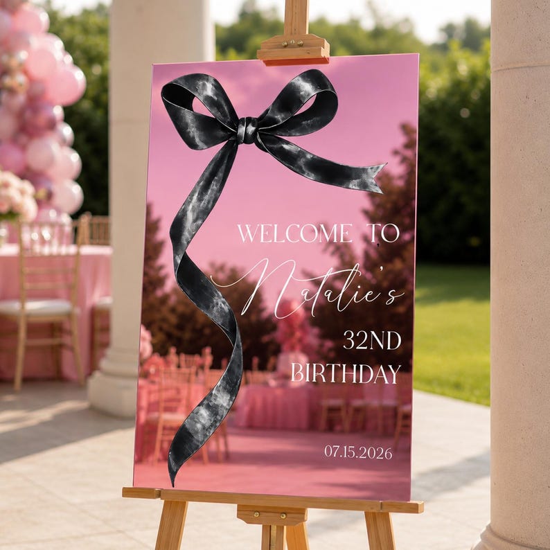 May include: A rectangular pink welcome sign on a wooden easel featuring a large black ribbon graphic. The text reads: WELCOME TO Natalie's 32ND BIRTHDAY 07.15.2026. The sign has a reflective surface showing a blurred outdoor event setting with pink decor.