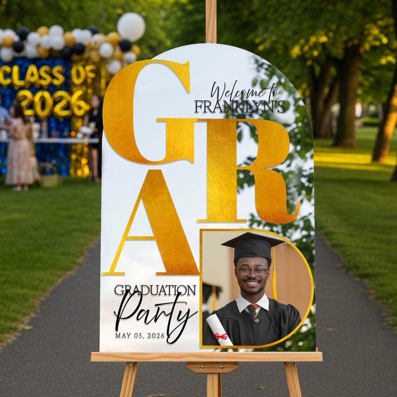 May include: A graduation party sign on an easel. The sign is white with gold lettering that spells out "GRAD" and "Graduation Party." It includes a photo of a graduate and the date, May 05, 2026. The sign welcomes guests to Franklyn's graduation.