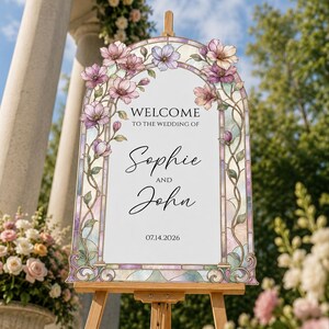 May include: An arched wedding welcome sign featuring a stained glass border with pink and purple floral illustrations. The text reads: WELCOME TO THE WEDDING OF Sophie AND John 14.07.2026. The sign is displayed on a wooden easel stand.