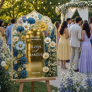 May include: An arched wedding welcome sign with a gold frame and center panel. The border features a floral design with blue, yellow, and white flowers. The text reads: WELCOME TO THE WEDDING OF OLIVIA and MARCUS JUNE 24, 2026. It stands on a wooden easel.