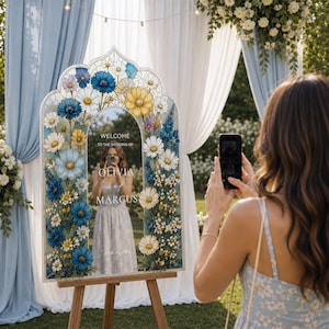 May include: An arched wedding welcome sign on a wooden easel featuring a floral border of blue, yellow, and white daisies. The center mirror displays the text: WELCOME TO THE WEDDING OF OLIVIA AND MARCUS JUNE 24, 202X in white lettering.