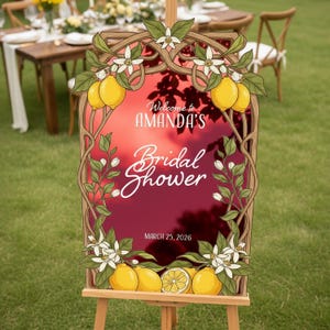 May include: A wooden easel displays a sign for Amanda's Bridal Shower, framed with lemon and floral illustrations. The sign features the text "Welcome to Amanda's Bridal Shower" and the date March 25, 2026, against a red sky backdrop.
