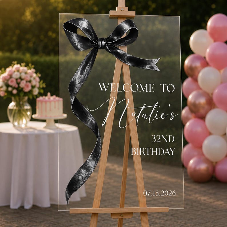 May include: A clear acrylic rectangular sign mounted on a wooden easel, featuring a large black and white marbled ribbon graphic at the top. The text reads: WELCOME TO Natalie's 32ND BIRTHDAY 07.15.2026 in white lettering with a script font for the name.