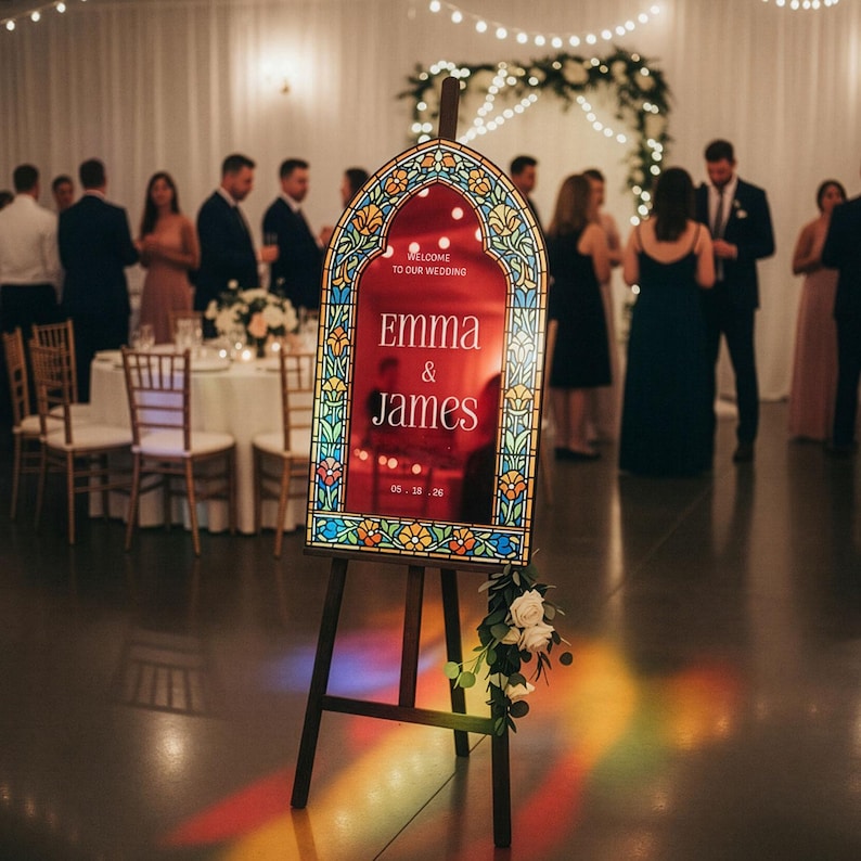 May include: An arched wedding welcome sign on a black easel featuring a red centre panel with the white text "WELCOME TO OUR WEDDING", "EMMA & JAMES", and "05.18.26". The frame has a colourful stained glass pattern with floral motifs and white roses.