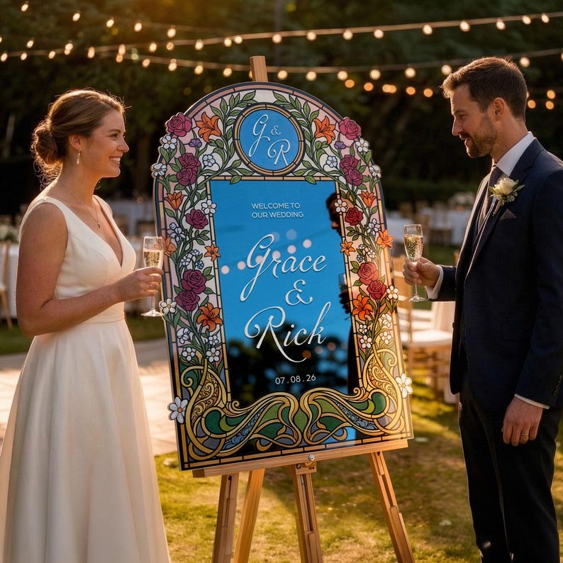 K&ouml;nnte beinhalten: Ein dekoratives Hochzeitsschild mit einer Spiegeloberfl&auml;che, umrahmt von bunten floralen Designs. Auf dem Schild steht "Welcome to Our Wedding" sowie die Namen "Grace & Rich" und das Datum 07.08.26. Das Schild steht auf einer Holzstaffelei.