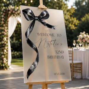 May include: A rectangular frosted acrylic welcome sign on a wooden easel, featuring a large black decorative ribbon bow on the left side. The text reads: WELCOME TO Natalie's 32ND BIRTHDAY 07.15.2026 in black lettering, centered on the smooth surface.