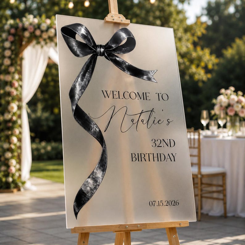 May include: A rectangular frosted acrylic welcome sign on a wooden easel, featuring a large black decorative ribbon bow on the left side. The text reads: WELCOME TO Natalie's 32ND BIRTHDAY 07.15.2026 in black lettering, centered on the smooth surface.