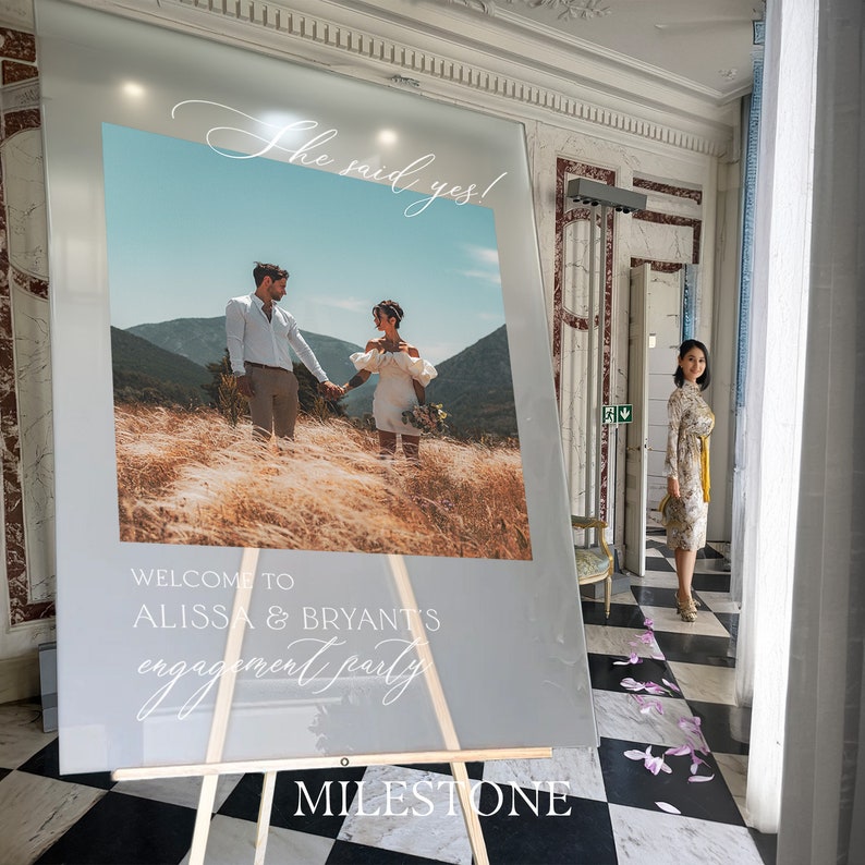May include: A rectangular frosted acrylic welcome sign on a wooden easel featuring a photograph of a couple in a field. The sign displays the text "She said yes!", "WELCOME TO ALISSA & BRYANT'S engagement party", and the word "MILESTONE" at the bottom.