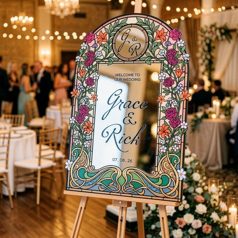 K&ouml;nnte beinhalten: Dekorativer Spiegel mit floralen Akzenten und den Namen "Grace & Rick" auf einer Staffelei. Der Spiegel ist mit bunten Blumen und Bl&auml;ttern umrahmt, mit dem Datum 07.08.26. Die Worte "Welcome to our wedding" stehen oben.